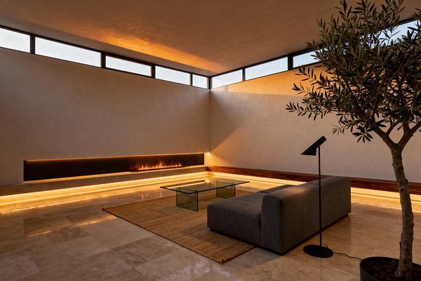 A minimalist living room with travertine floors and smooth plaster walls centers on a long recessed linear fireplace, its warm LED lighting extending around a perimeter plinth. A low gray sofa faces a clear glass coffee table on a textured jute rug, paired with a slim black floor lamp. Clerestory windows wash the room with soft daylight, and a potted indoor tree adds greenery, emphasizing clean lines, negative space, and a calm, modern mood.