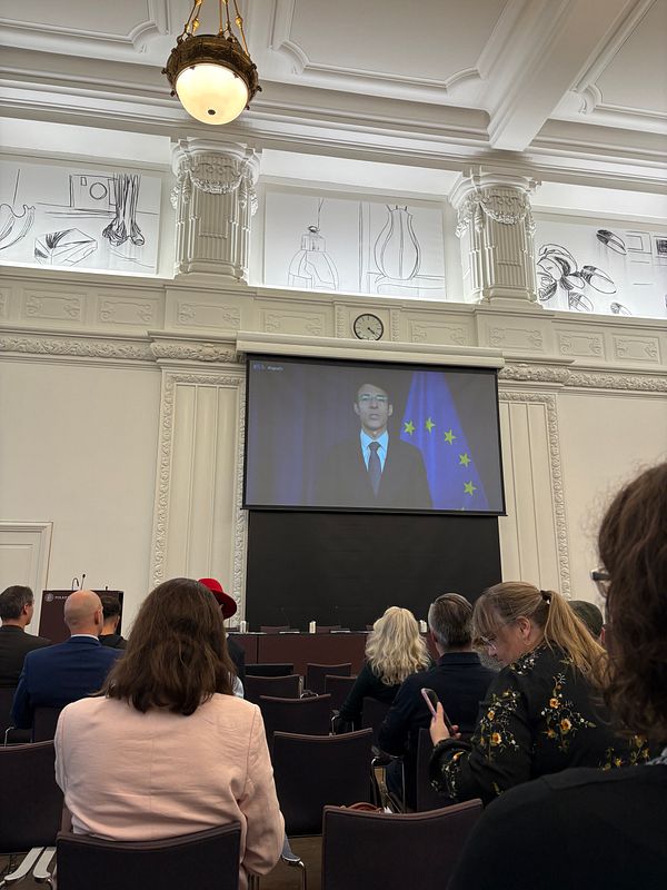 A conference setting with a speaker displayed on a large screen and attendees seated in the foreground.