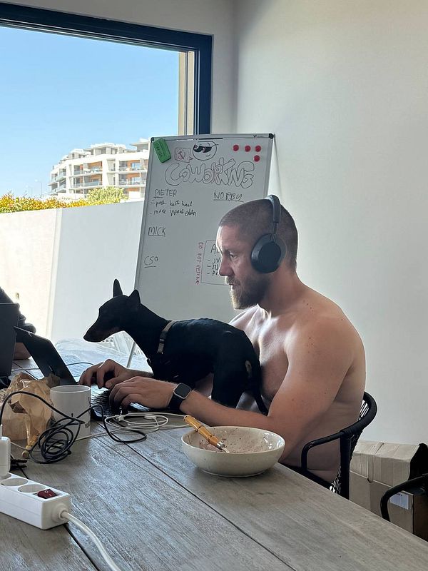A man is working on a laptop at a table with a small dog on his lap, in a bright, modern workspace.