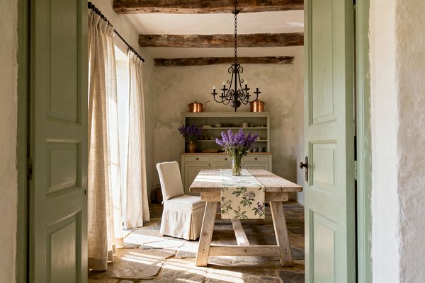 A cozy French Country dining room framed by pale green doors, featuring a weathered timber trestle table with a floral runner and a glass vase of lavender. A slipcovered linen chair sits beside the table, while a sage-green hutch with copper pots anchors the back wall. Exposed wood ceiling beams, textured plaster walls, stone flagstone floors, and an ornate black wrought-iron chandelier complete the softly sunlit space with natural linen curtains.