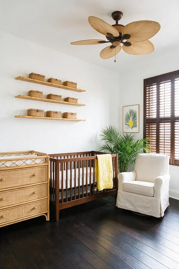 A bright nursery with white walls, dark wood floors, and tropical-inspired details. The room includes a wooden crib, a rattan dresser, floating bamboo shelves with baskets, a cream slipcovered chair, a palm plant, shuttered windows, and a ceiling fan with leaf-shaped blades.