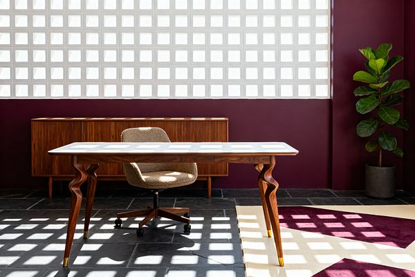 Mid-century inspired home office with a deep burgundy wall and a large white breeze-block window casting a checkerboard of light across a dark slate floor. A walnut desk with a white top and sculptural legs with brass caps sits center stage, paired with a taupe boucle swivel chair on casters. A ribbed walnut credenza runs along the back wall, and a fiddle-leaf fig in a charcoal planter softens the corner. A cream-and-plum geometric rug adds color under the desk.