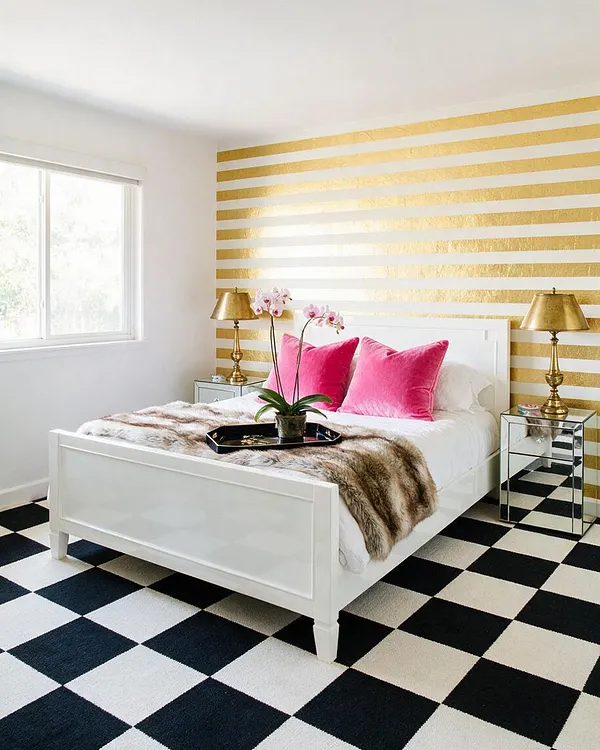 A bright master bedroom with a white bed centered against a gold-and-white striped accent wall. The bed is dressed in white bedding, hot pink velvet pillows, a faux fur throw, and matching mirrored nightstands with gold table lamps, all set over a bold black-and-white checkerboard floor.