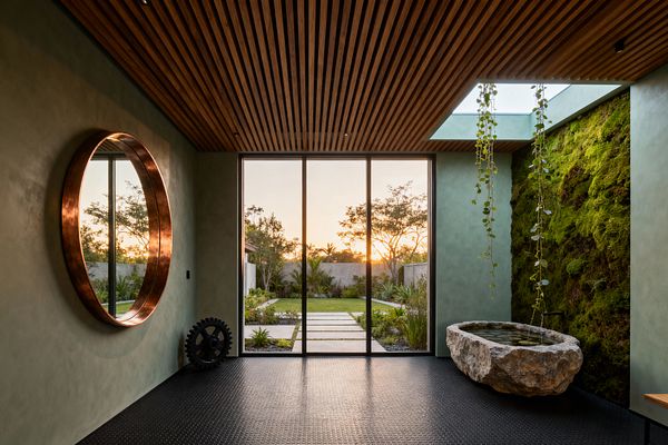 A serene home gym with pale green plaster walls features a large round copper-framed mirror, floor-to-ceiling glass doors opening to a garden, and a slatted wood ceiling with a skylight. A living moss wall with trailing vines borders a rough-hewn stone water basin, while black coin-pattern rubber flooring and minimal equipment, including a gear-shaped weight, create a calm, tactile space flooded with warm natural light.