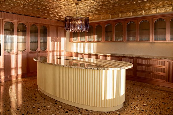 A vintage glam kitchen with an antique gold pressed-metal ceiling, an amethyst crystal chandelier, and long runs of cherry cabinetry featuring fluted fronts and arched reeded-glass doors. A curved island with a ribbed cream base and beige granite top anchors the room, while warm under-cabinet lighting and a speckled terrazzo floor enhance the soft, retro elegance.