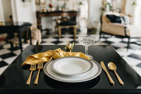 A glamorous dining vignette features a glossy black table set with stacked white plates edged in Greek key detail, gold flatware, a folded mustard-gold napkin, and a crystal coupe glass. In the softly blurred background, a black-and-white checkered floor, light upholstered seating, and reflective decor reinforce the elegant Hollywood Regency atmosphere.