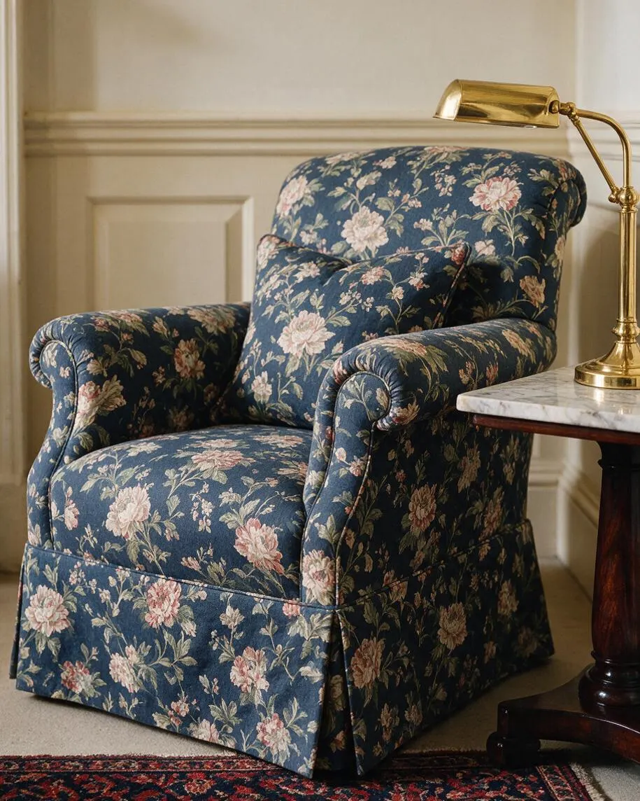 A traditional upholstered armchair in dark blue floral fabric sits beside a marble-topped side table with a polished brass desk lamp. The chair has rolled arms, a plush cushion, and a skirted base, set against cream paneled walls and a patterned rug.