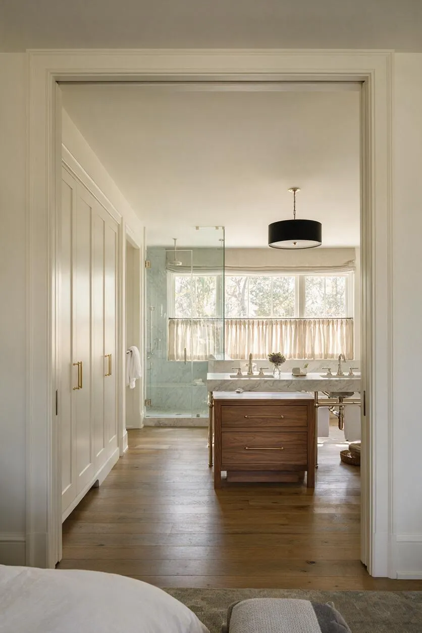 A transitional master bathroom viewed through a wide doorway, featuring a warm wood vanity with a marble countertop, brass fixtures, and a large window dressed with a light curtain. A glass-enclosed shower sits to the left, and white built-in closet doors line the entry wall.
