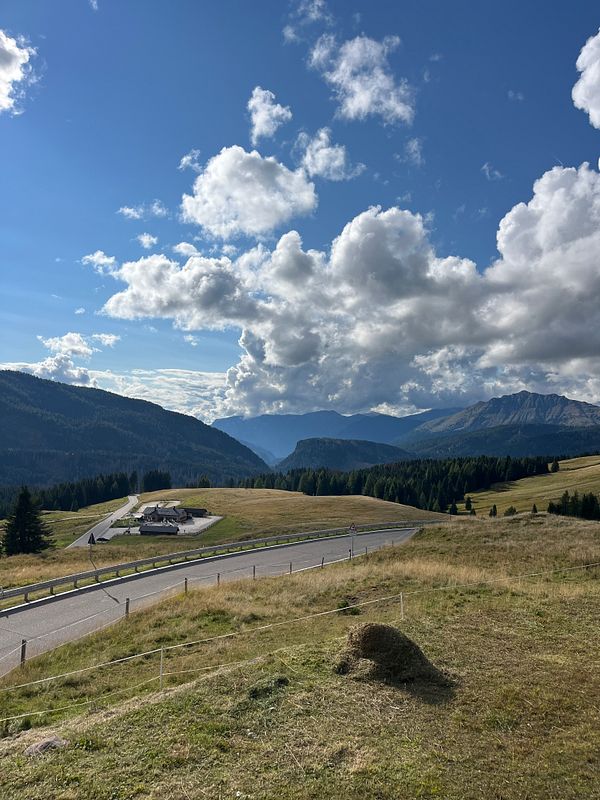 A scenic view of the Dolomites featuring rolling hills, a winding road, and dramatic cloud formations.