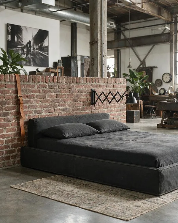 A moody industrial bedroom features a low black platform bed against a short exposed brick wall, with two dark pillows and a minimalist black wall lamp. The space includes concrete flooring, a muted woven rug, metal columns, exposed ductwork, and vintage-style furnishings in the background.