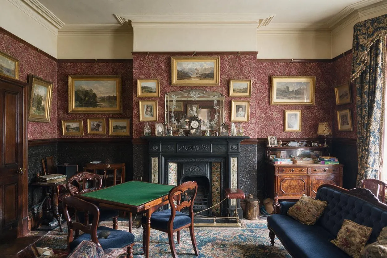 A Victorian-style game room with dark patterned wallpaper, black carved fireplace, and a green felt card table surrounded by wooden chairs. The room includes a navy tufted sofa, ornate area rug, gilt-framed paintings, and heavy patterned drapes by the window.