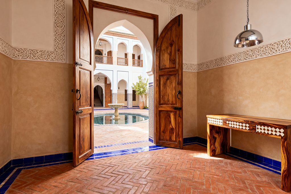A Moroccan-style home office features a slim wooden desk with white diamond inlay beneath a chrome dome pendant. Double arched wooden doors open to a bright courtyard with a tiled fountain and carved arches. Walls are sand-colored lime plaster with a white carved frieze, and the floor is terracotta herringbone brick edged in cobalt-blue tiles. Wrought-iron door hardware and rich wood grain create a warm, handcrafted atmosphere.