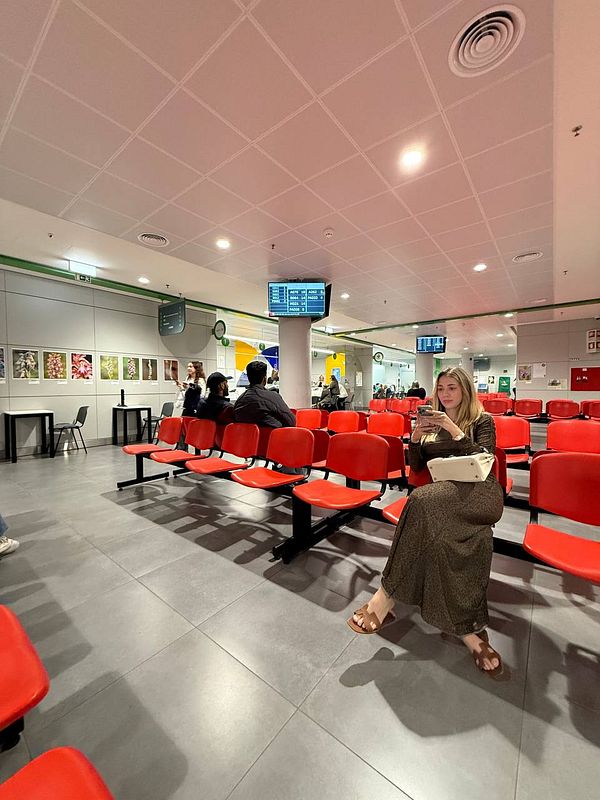 A woman sits in a waiting area of a driver license center surrounded by red chairs and other visitors.