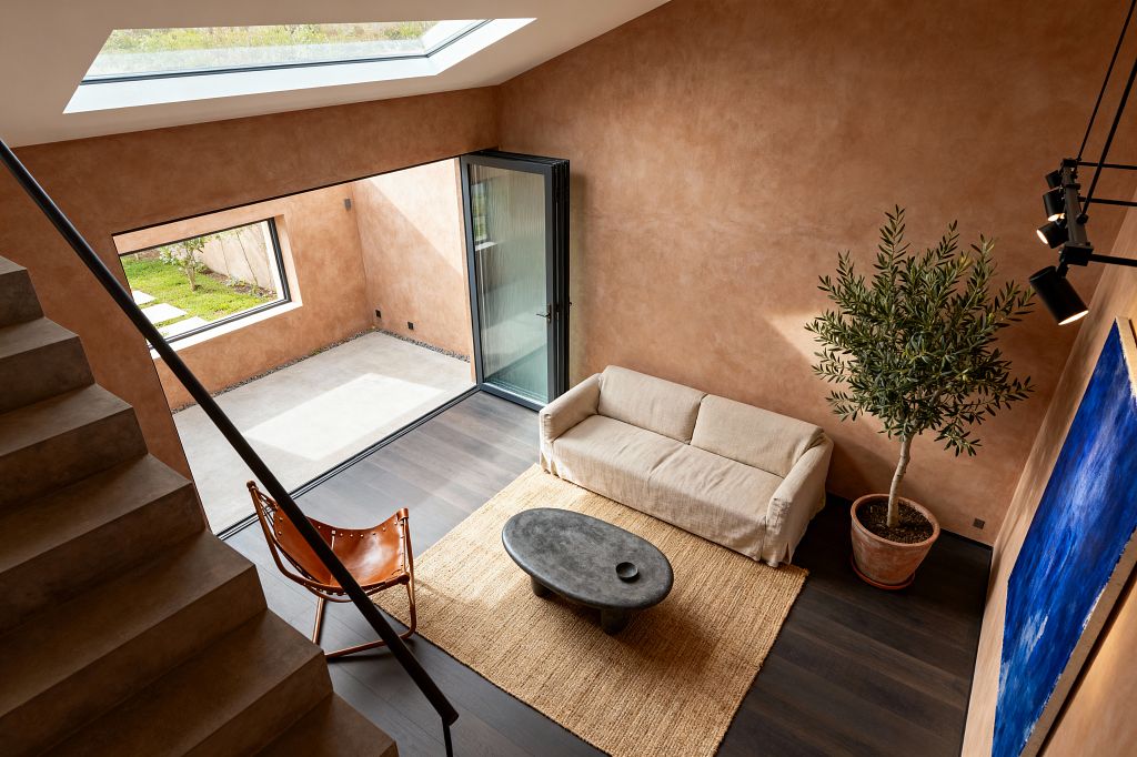A high-ceilinged contemporary living room with warm terracotta plaster walls, a large roof skylight, and black-framed folding glass doors opening to a small courtyard. Dark wood floors support a jute rug, beige linen sofa, oval charcoal concrete coffee table, and a cognac leather sling chair. A potted olive tree in a terracotta planter sits beside a bold blue canvas, while black track spotlights and a minimalist stair with a dark metal rail complete the space.