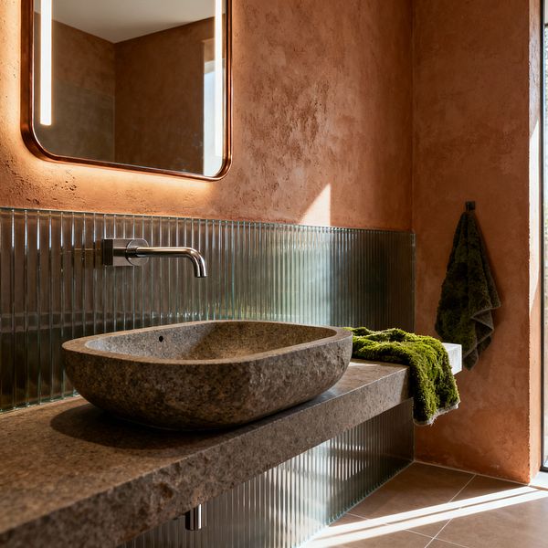 A contemporary bathroom features terracotta plaster walls, a floating gray natural-stone vanity with a carved oval vessel basin, and a wall-mounted stainless steel spout beneath a rounded-corner mirror with integrated vertical lights. A ribbed fluted glass backsplash lines the sink wall, sunlight enters from the right casting long shadows across taupe floor tiles, and a moss-green towel is draped on the counter with another hanging on a hook.