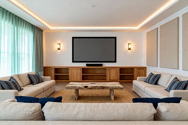 A coastal-inspired media room features three beige linen sofas arranged in a U around a rustic driftwood coffee table atop a seagrass rug. A large flat-screen and soundbar mount on a white shiplap wall above a long natural-wood built-in with open shelves and sliding doors, flanked by warm ribbed-glass sconces; sea-glass aqua curtains line the windowed left wall, sand-colored acoustic panels hang to the right, light wood flooring and ceiling cove LEDs create a soft, inviting glow.