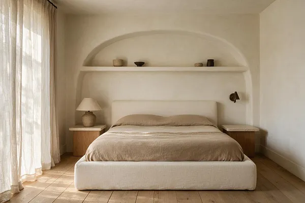 A calm guest bedroom with a low upholstered bed centered in a soft arched wall niche. Linen curtains filter daylight at the left, while wood nightstands, ceramic lamps, and small decorative vessels add warm natural texture.