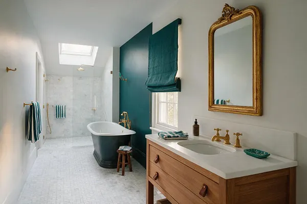 A spacious master bathroom with white tiled floors and shower walls, a dark freestanding bathtub, and a natural wood vanity topped with a white countertop. The room features a deep teal accent wall with matching window shade, brass fixtures and mirror, skylight, and teal striped towels for a layered eclectic look.