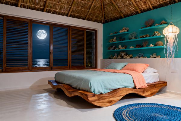 A tropical-themed kids’ bedroom at night with a sculptural wave-shaped wooden platform bed dressed in seafoam bedding and a coral-pink throw. A turquoise accent wall behind the bed has four floating shelves displaying coral and sea-life decor. A jellyfish-style paper pendant glows to the right. The ceiling is palm thatch, the floor is polished light concrete with a round braided blue rug, and wooden louvered windows reveal a full moon over the ocean.
