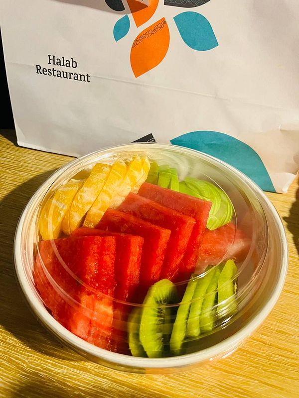 A colorful fruit platter is displayed in a clear container next to a restaurant bag.