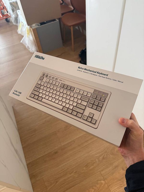A person is holding a boxed 8BitDo retro mechanical keyboard in a home setting.