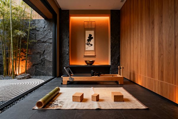 A serene home gym features a light-wood Pilates reformer before a backlit alcove holding a sumi-e scroll and bronze bowl; a beige mat with cork yoga blocks rests on charcoal slate tiles. To the left, floor-to-ceiling glass opens to a zen garden with raked white gravel, river stones, and bamboo, while a rugged stone wall and warm oak paneling with concealed uplighting create a calm, minimalist setting.