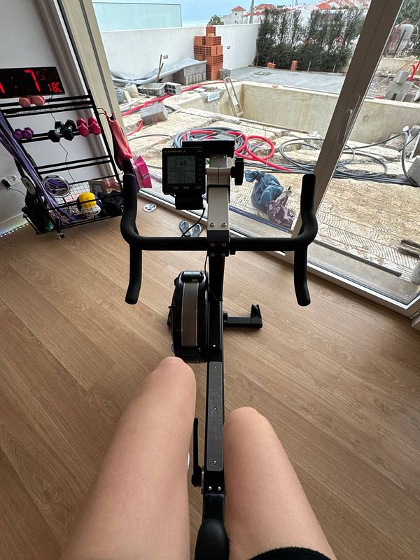 A person is seated on a rowing machine, with a view of a construction site through a large window.