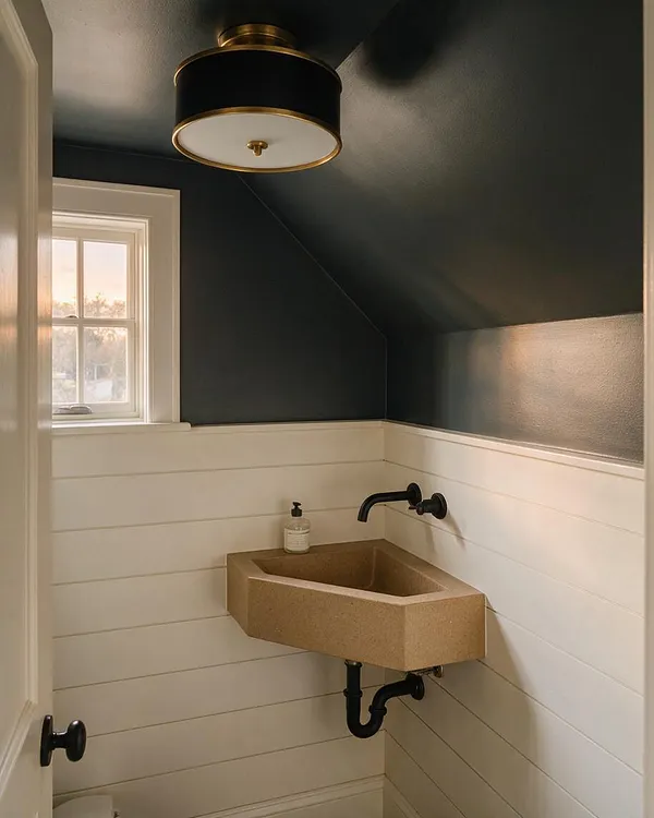 A compact powder room with dark charcoal upper walls and white horizontal shiplap below, centered around a small faceted stone sink with matte black wall-mounted faucet and exposed black plumbing. A round flush-mount ceiling light with brass trim and a small window with white trim add warmth and softness.