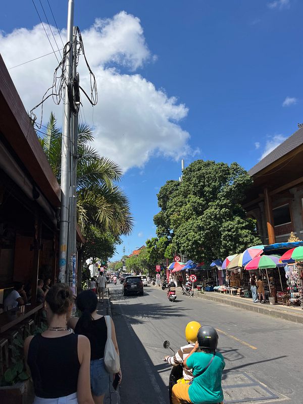 A bustling street scene in Ubud, showcasing shops, pedestrians, and vibrant market stalls under a clear blue sky.