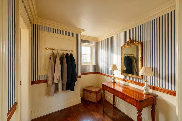 A traditional entryway with blue-and-white striped wallpaper, cream trim, and polished wood flooring. A mahogany console table with two table lamps sits beneath a gilded wall mirror, with a coat rack, woven storage chest, and small window adding function and charm.