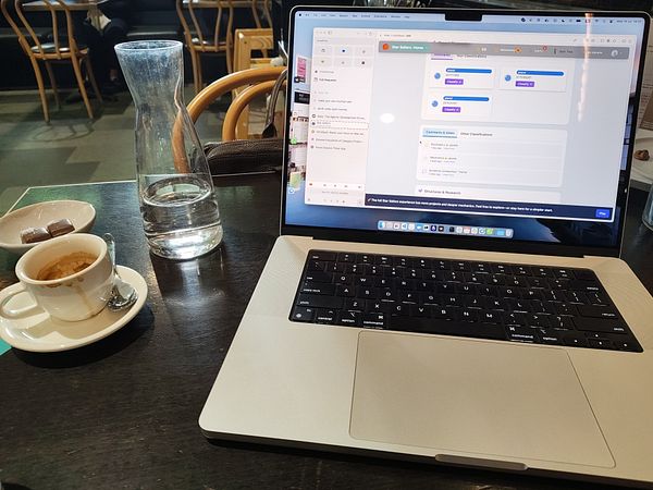 A laptop is set up on a table in a café, displaying a project management interface alongside a cup of coffee and a carafe of water.