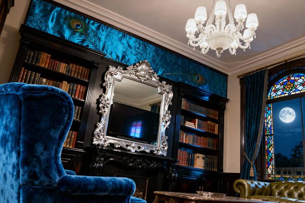 A richly detailed Victorian media room features ebonized bookcases and a carved fireplace with a TV framed by an ornate silver mirror. A sapphire blue velvet wingback chair, olive tufted leather sofa, marble-topped table, and deep teal drapes flank an arched stained-glass window, while peacock-feather silk damask panels and a white multi-arm chandelier add shine and warmth to the moody, jewel-toned space.