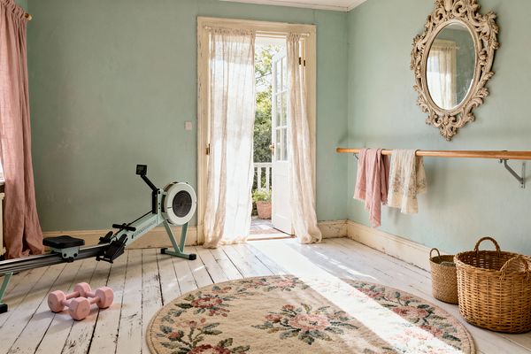Light-filled shabby chic home gym with seafoam walls and distressed white plank floors. French doors with gauzy linen curtains open to a garden. A wooden ballet barre spans the wall beneath an ornate antiqued oval mirror, with pink and cream cloths draped over it. A mint rowing machine and pastel pink dumbbells sit near a round floral rug, alongside woven baskets. Soft daylight creates an airy, romantic atmosphere.