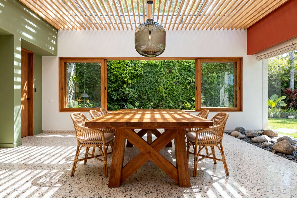 A sunlit dining room features a thick solid-wood table and six rattan chairs on speckled terrazzo flooring. A skylight with timber slats casts striped shadows, a smoked-glass pendant hangs centrally, large wood-framed windows face a lush living wall and garden, and a river-stone border lines the edge beside white plaster, a sage-painted wall, and a terracotta soffit.