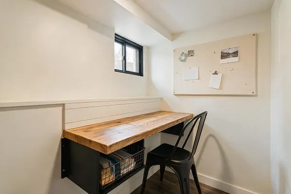 A small farmhouse-style basement workspace with a live-edge wood desk, white painted walls, a narrow black-framed window, and a beige bulletin board on the wall. A black metal chair sits at the desk, which has open storage underneath and simple, clean finishes.