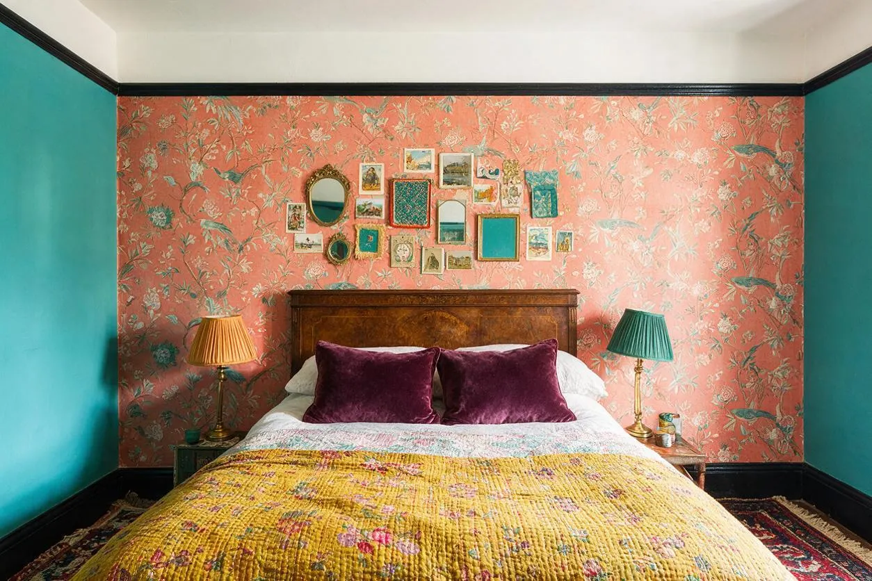 A maximalist guest bedroom features a wooden bed centered against coral floral wallpaper, with a curated cluster of small framed pictures and mirrors above the headboard. Teal painted side walls, a mustard quilt, plum velvet pillows, and mismatched lamps in warm gold and teal create a bold, layered look.