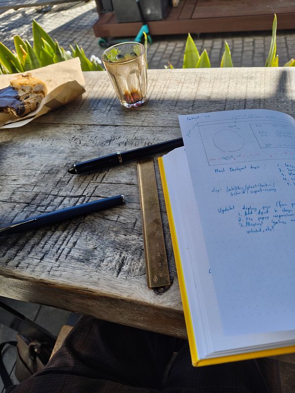 A workspace setup featuring a notebook, pens, a glass, and a cookie on a wooden table.