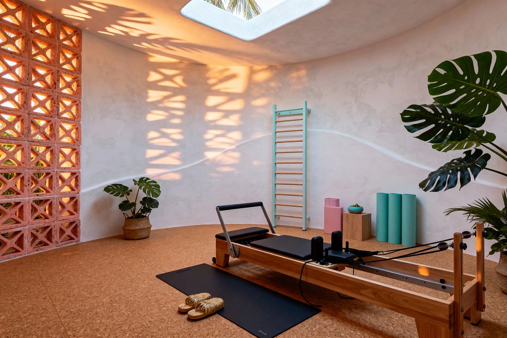 Sunlit home gym with a wood Pilates reformer on cork flooring sits below a rectangular skylight where palm fronds are visible, while a coral terracotta breeze-block wall casts patterned shadows on warm white plaster. Mint wall bars, seafoam rollers, a pastel pink block, a small wood pedestal, a black mat with straw slippers, and potted monstera and palm plants complete the space.