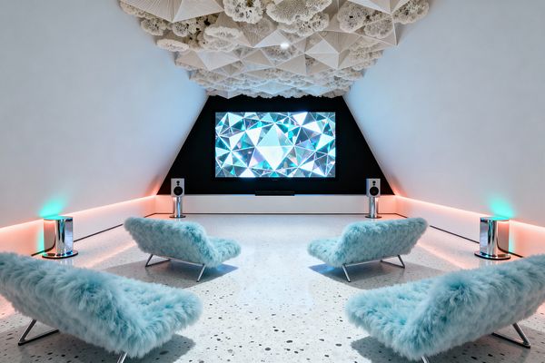 Futuristic media room with a large screen mounted in a matte black triangular wall, viewed from behind four low lounge chairs upholstered in icy blue faux fur on chrome sled legs. The white terrazzo floor and sloped white walls are edged with pink and teal LED cove lighting. Two white speakers on chrome pedestals stand beside the screen, with glossy cylindrical side tables near the walls. Above, a sculpted origami-style ceiling and cloud-like acoustic panels add texture.