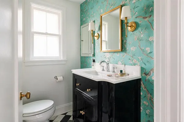 A compact powder room features turquoise floral wallpaper on one wall, a gold-framed mirror, and brass wall sconces with white shades. Below, a glossy black vanity with a white countertop and chrome faucet sits beside a white toilet, with black-and-white checkered flooring adding contrast.