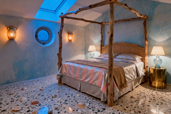 A coastal bedroom with a rustic driftwood four-poster bed and woven rattan headboard, dressed in silvery satin bedding and a tan jute throw. The terrazzo floor is scattered with seashells. Soft blue, plaster-washed walls feature two amphora-style sconces and a round porthole window. A ceiling skylight casts cool blue light, while glass table lamps with beige shades sit beside the bed, one on a polished brass drum nightstand, creating a warm nautical atmosphere.