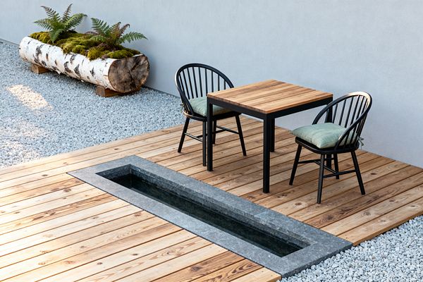 A minimalist Scandinavian patio features a light pine wood deck with a narrow rectangular water rill lined in gray stone. A small square table with a solid-wood top and black metal frame is paired with two black spindle-back chairs fitted with pale green cushions. Surrounding the deck is light gray gravel and a smooth pale wall. A hollow birch log planter filled with moss and ferns sits along the edge, adding natural texture.