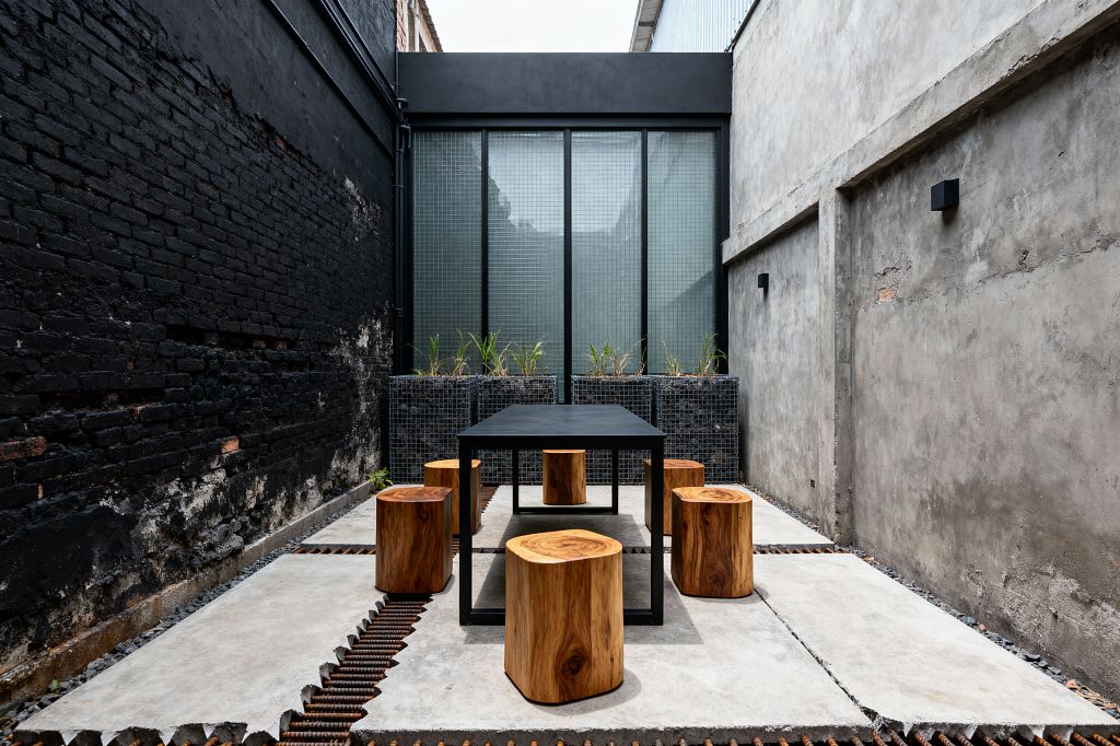A narrow industrial patio features cast-concrete slabs separated by exposed steel rebar channels. A matte black metal dining table sits centrally, surrounded by six solid wood log stools. One wall is weathered black brick and the opposite wall is raw gray plaster with black cube sconces. At the far end, three gabion planters filled with dark stones and grasses stand before tall wired-glass panels in a black frame, providing diffused daylight.