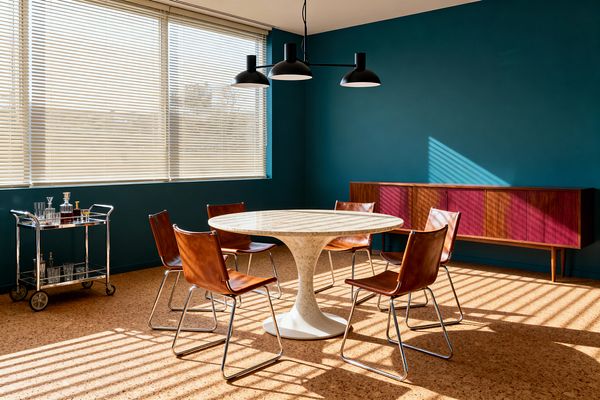 A mid-century modern dining room features a round white pedestal table with a light marble top, surrounded by six cognac leather sling chairs on chrome cantilever frames. A black three-shade pendant hangs above, while deep teal walls, a long walnut credenza with magenta sliding doors, and a chrome-and-glass bar cart define the space; cork flooring and wide windows with cream blinds cast striped sunlight across the room.