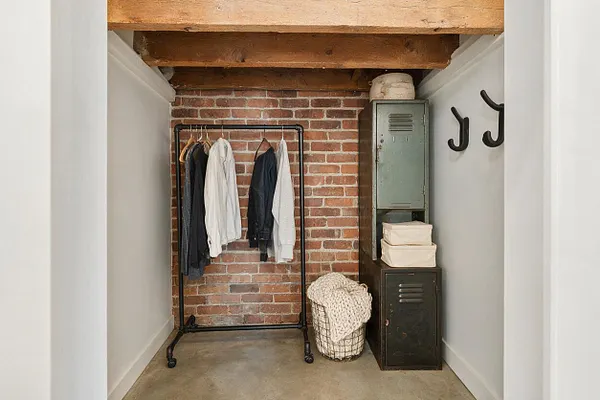 A narrow closet features exposed brick at the back, thick rustic wood beams overhead, and white side walls. A black metal clothing rack holds a few hanging garments, while stacked vintage-style lockers, wall hooks, and a woven basket add storage and texture.