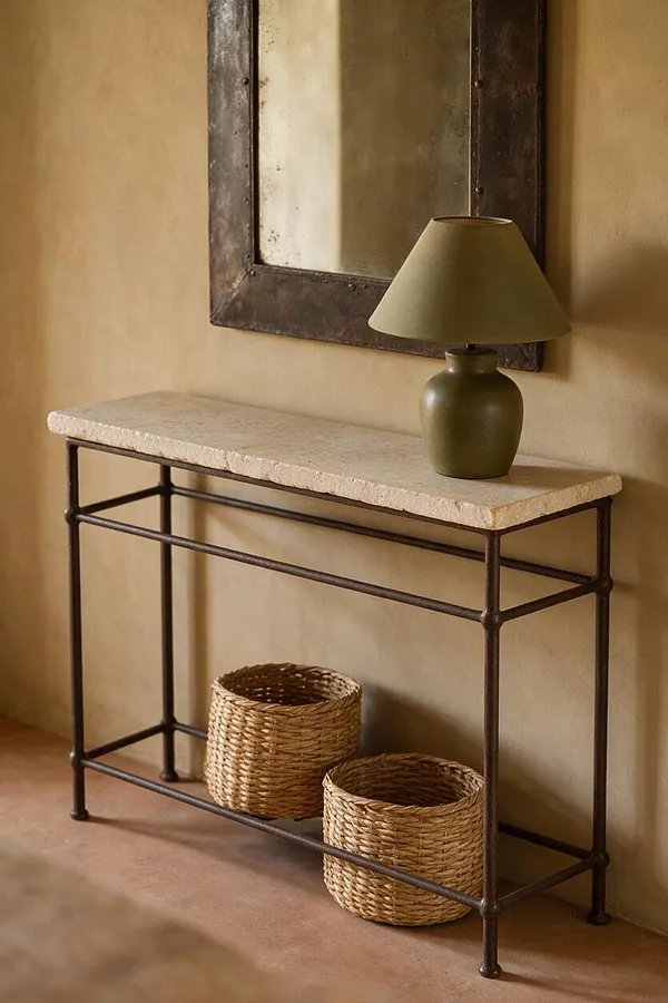 A Mediterranean-style entry vignette with a slim console table topped by a pale stone slab and dark metal frame. Above it hangs a rustic mirror, and an olive-green table lamp sits on the right side of the console. Two woven basket planters or storage baskets rest on the floor beneath, against warm beige plaster walls and a terracotta-toned floor.