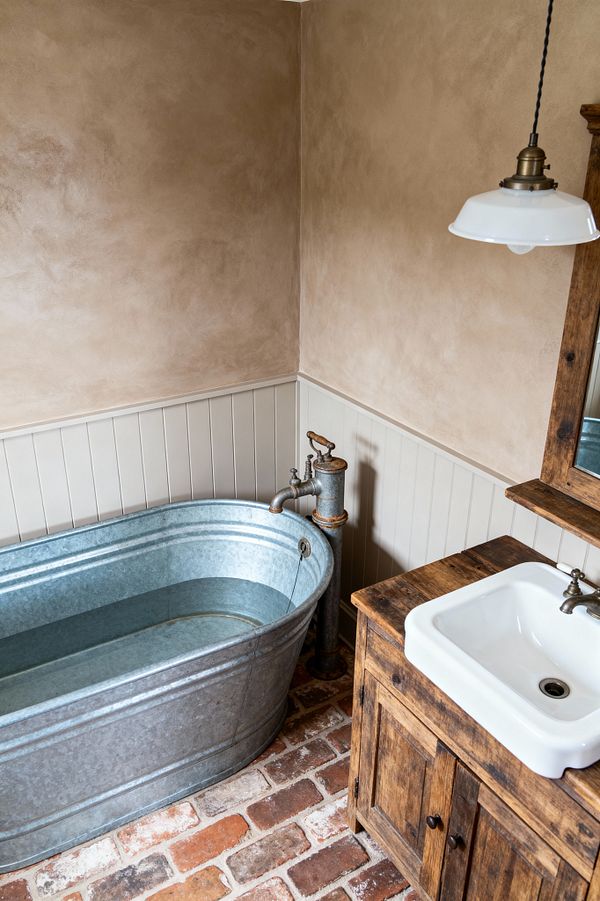 A farmhouse bathroom featuring a galvanized steel soaking tub beside a vintage hand-pump style filler, set against cream-painted beadboard and warm taupe plaster walls. The floor is reclaimed red brick, and a rustic wood vanity with a white ceramic sink and aged metal tap sits under a wood-framed mirror, while an enamel-shaded pendant light hangs above, creating an authentic, timeworn look.