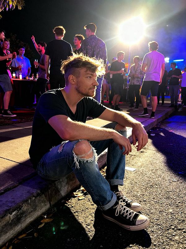 A young man sits on the curb at a lively outdoor party, surrounded by a crowd of people enjoying the night.