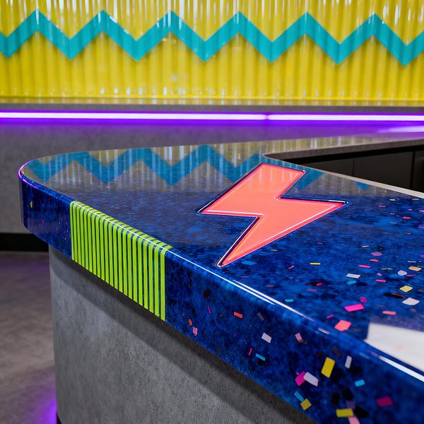 A Pop Art kitchen island with a glossy electric-blue resin countertop features a neon-coral lightning-bolt inlay and lime green pinstripes at the rounded edge. Multicolored confetti chips speckle the surface; a corrugated yellow backsplash carries a teal zigzag band, and purple LED strip lighting washes the base over concrete-look cabinetry.