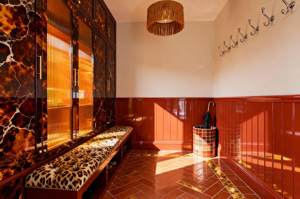 A maximalist mudroom features glossy oxblood wainscoting, a bank of tortoiseshell-lacquer cabinets with ribbed amber glass doors and brass pulls, and a built-in bench topped with a tufted leopard-print cushion. Sunlight falls across a terracotta herringbone tile floor with gold inlay borders, while a caramel fringe pendant, a line of polished chrome wall hooks, and a cylindrical red-and-white mosaic umbrella stand holding a black umbrella and cane complete the richly patterned, high-contrast space.